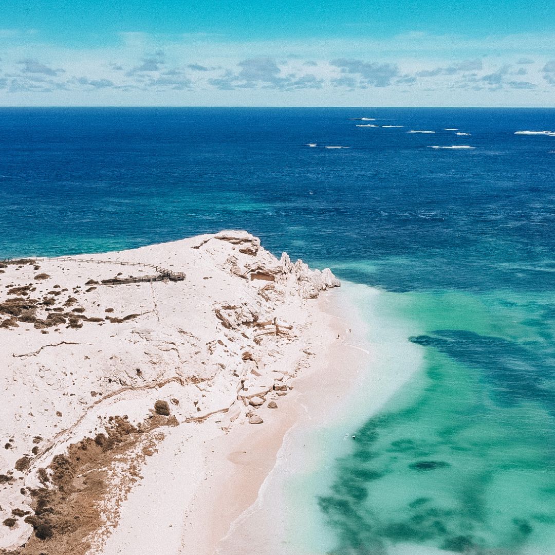 An aerial view of the white cliffs and the board walk and lookout on top. The cliffs edge is left to middle of the image and surrounded by beautiful blue ocean in shades ranging from light turquoise to deep blue