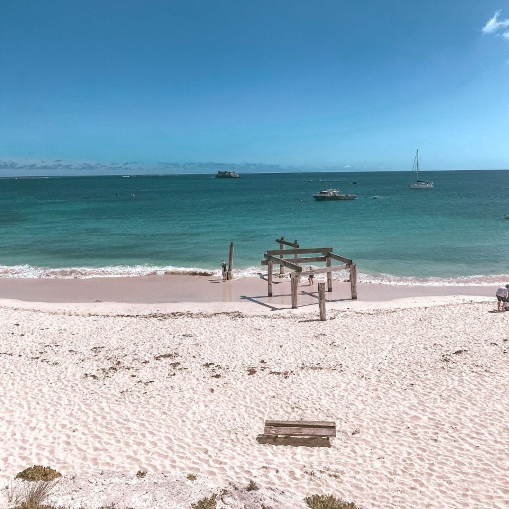 the white sand of Hamelin bay beach with a bench seat on the sand. in front of the bench are the old jetty pylons. beyond the jetty is he clear blue water of Hamelin Bay with a few boats floating in the ocean