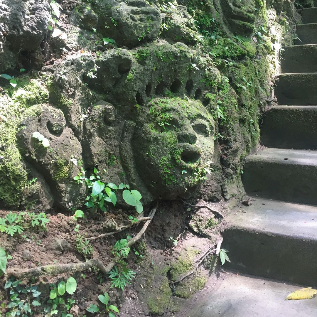 a craved face into the rock wall with stairs to the right of the image. the carved face is covered in green moss