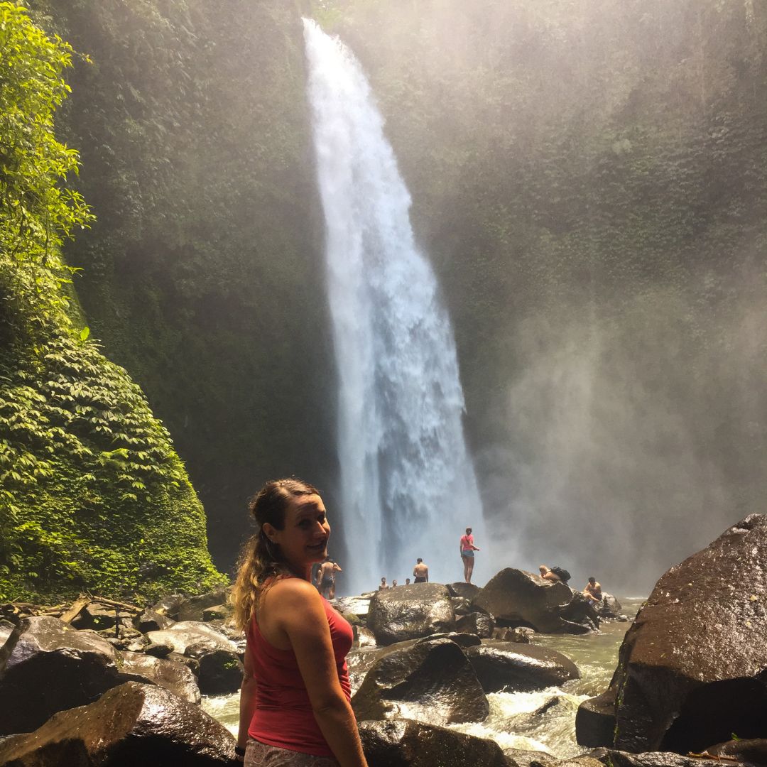 Luisa is standing in front of Nungnung waterfall but looking back toward the camera looking over her right shoulder. She is wearing a red tank top. The waterfall is beyond her in the middle of the image. there a few people standing on the boulders at the base of the waterfall