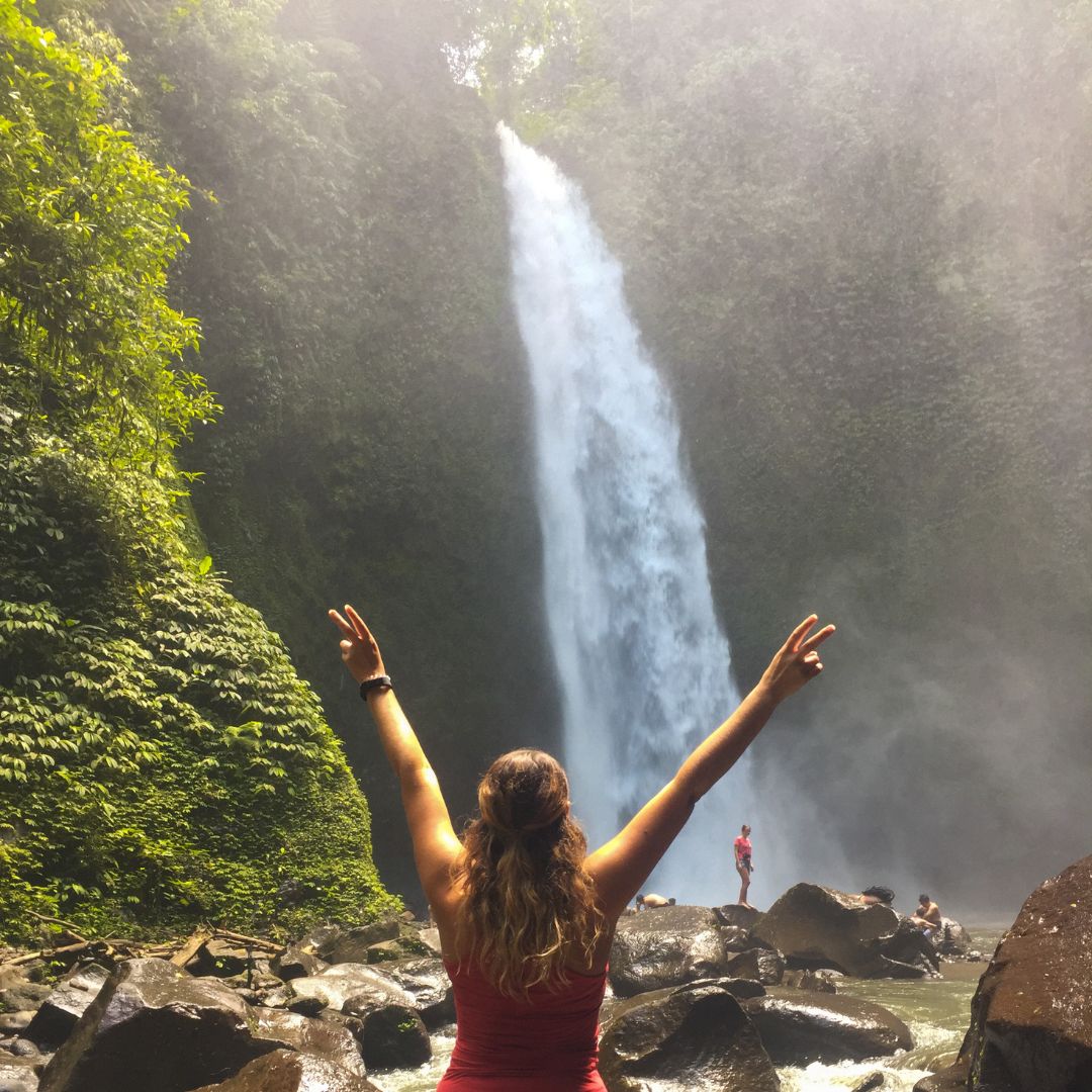 Luisa is standing in front of the waterfall and looking toward the falls. She has both arms raised above her head and giving the peace sign with her fingers. She is wearing a red tank top. The waterfall is beyond her in the middle of the image. there a few people standing on the boulders at the base of the waterfall