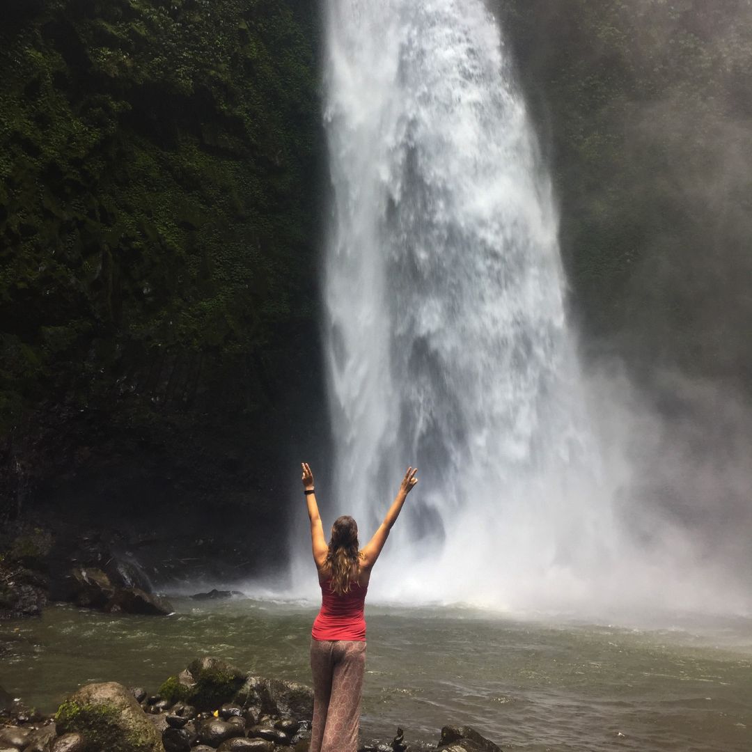 Luisa is close to the base of the waterfall. She is watching the waterfall while facing away from the camera. She has both arms raised and her hands in the peace symbol. She is wearing a red singlet top and brown pants