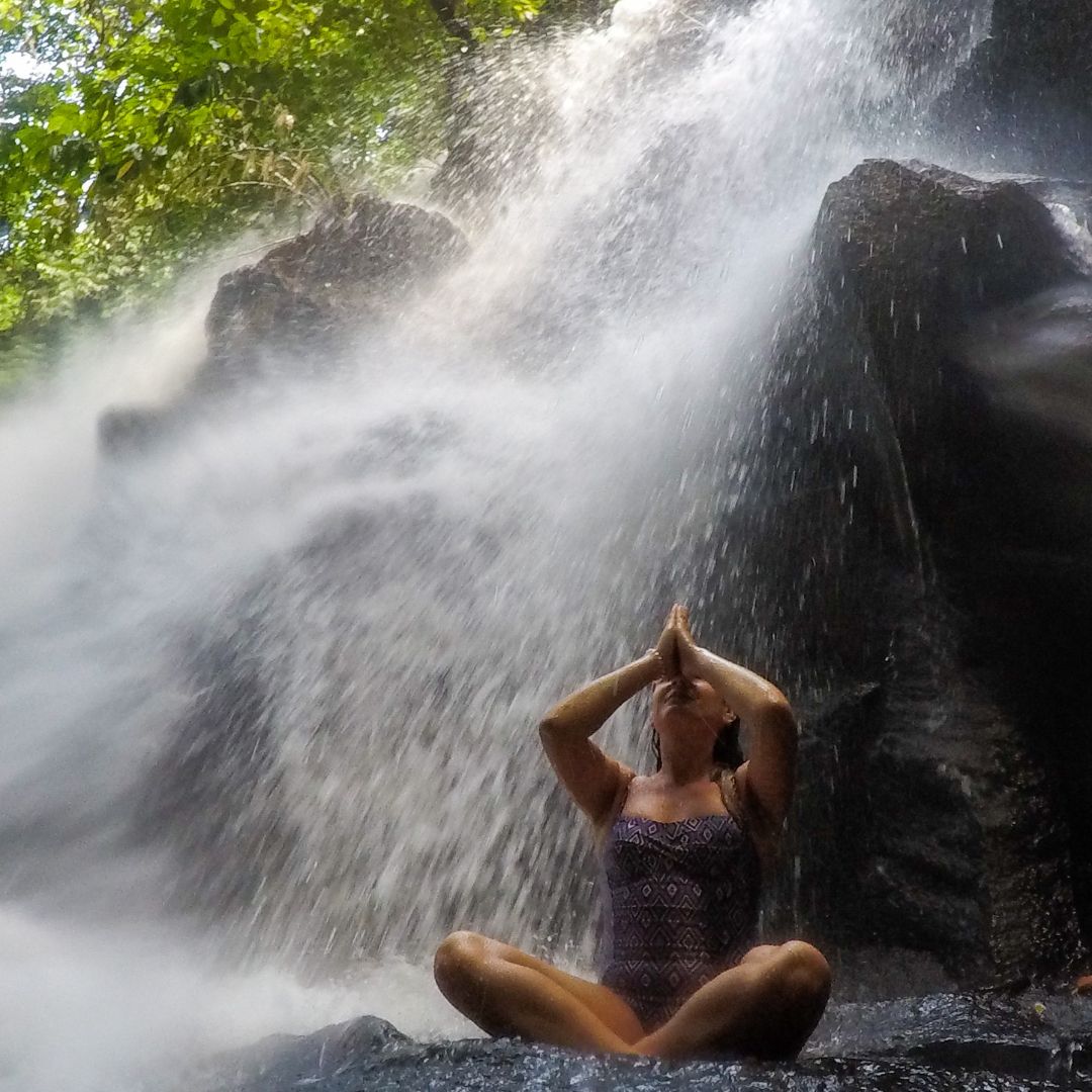 Luisa sitting at the base of the waterfall under the water. She has her legs crossed and her arms raised with her hands in front of her face in the namaste pose.