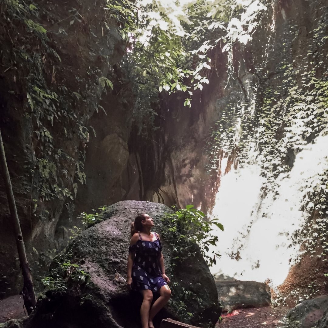 Luisa is leaning on a boulder and looking up and over her left shoulder (right of the image) She is in the save, surrounded by the rock walls and green jungle falling over the sides. there is sunlight coming down the right of the image