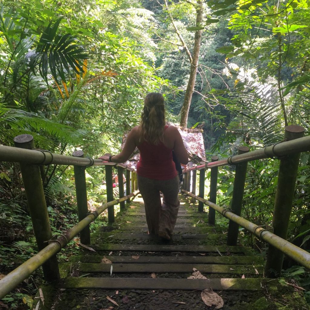 Luisa is walking down some mud and bamboo stairs towards the waterfall. she is facing away from the camera as she walks down and has her left hand on the hand rail. she is wearing a red singlet top and brown pants. the stairs have bamboo rails on either side and there is lush green jungle surrounding her and the stairs