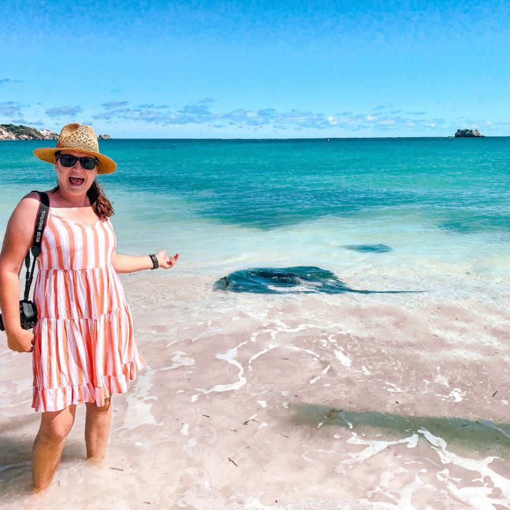 Luisa is standing ankle deep in the clear blue water. There are two stingrays in the water behind her. one large and one small sting ray with the larger one closer to Luisa. She is wearing an orange and white striped summer dress and straw hat. she has her canon camer slung over her right shoulder and is pointing to the stingrays with her left hand. She is so excited she has her mouth open in a huge smile