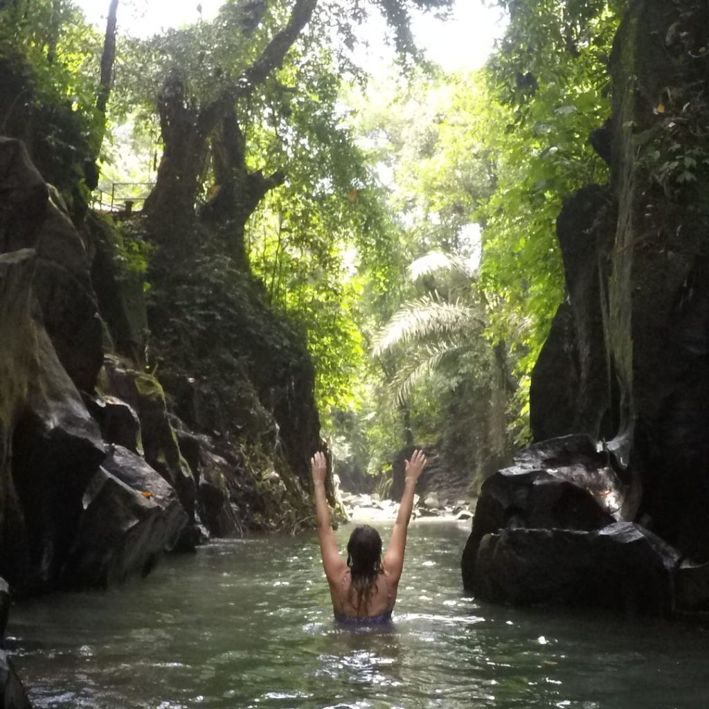 Luisa is standing in the hidden river at Kanto Lampo Waterfall. She is waist deep standing in the water and has both arms raised above her head. she is looking away from the camera down the river canyon toward lush green jungle. there are tall rock faces on either side of the river