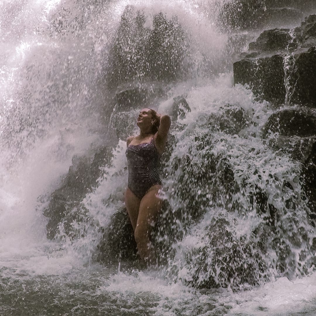 Luisa standing at the base of the waterfall under the water. She is posing with her left arm behind her head and her face facing upward.