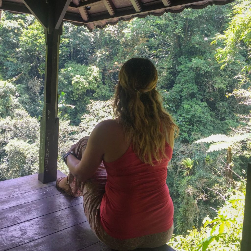 Luisa is sitting in one of the rest stops found along the stairs. She is sitting on the wooden floor with her left leg bent up and her left arm holding her leg. She is looking out into the green jungle in front of her