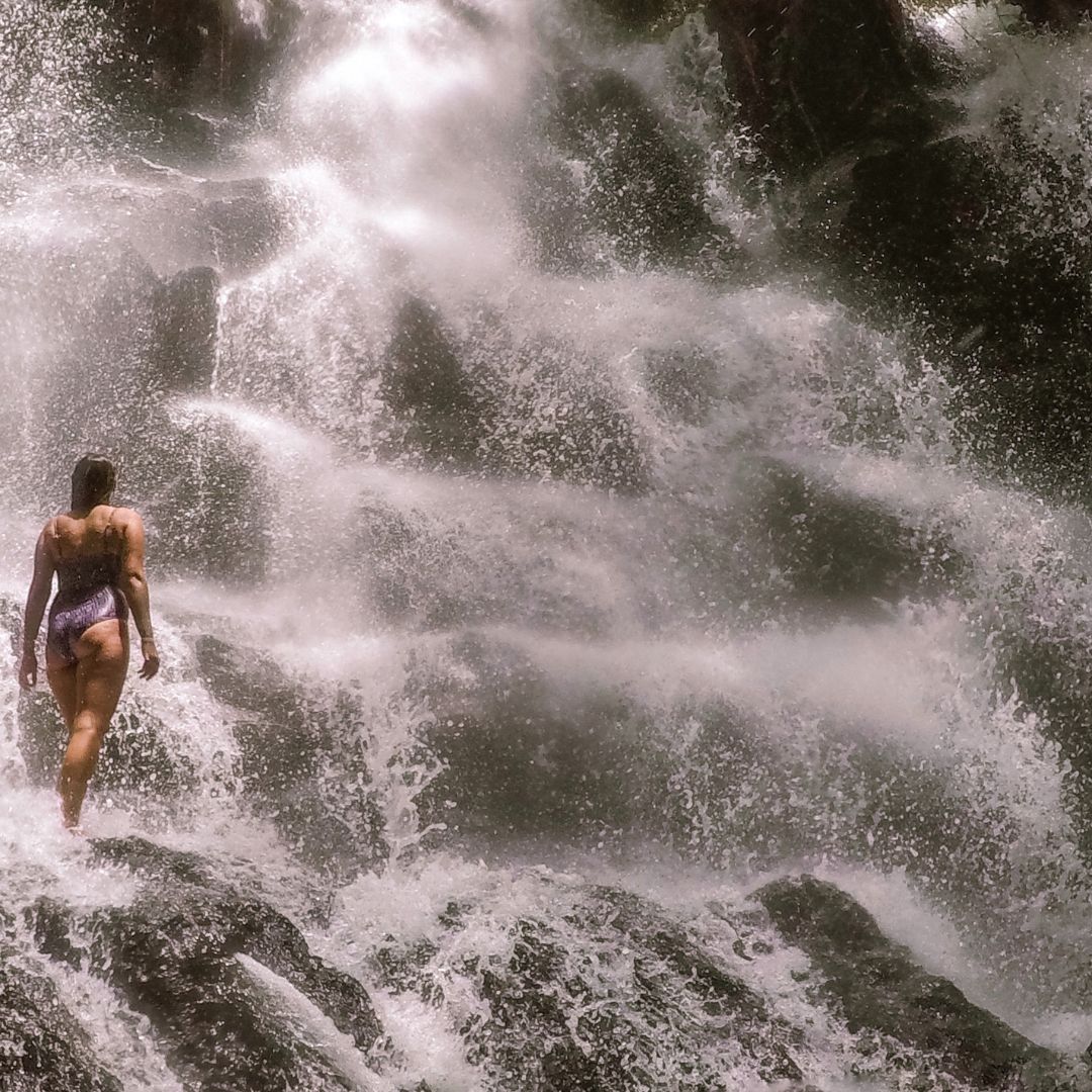 Luisa is standing on the rocks under the waterfall to the left of the image. she is wearing purple bathers and facing away from the camera and looking at the waterfall