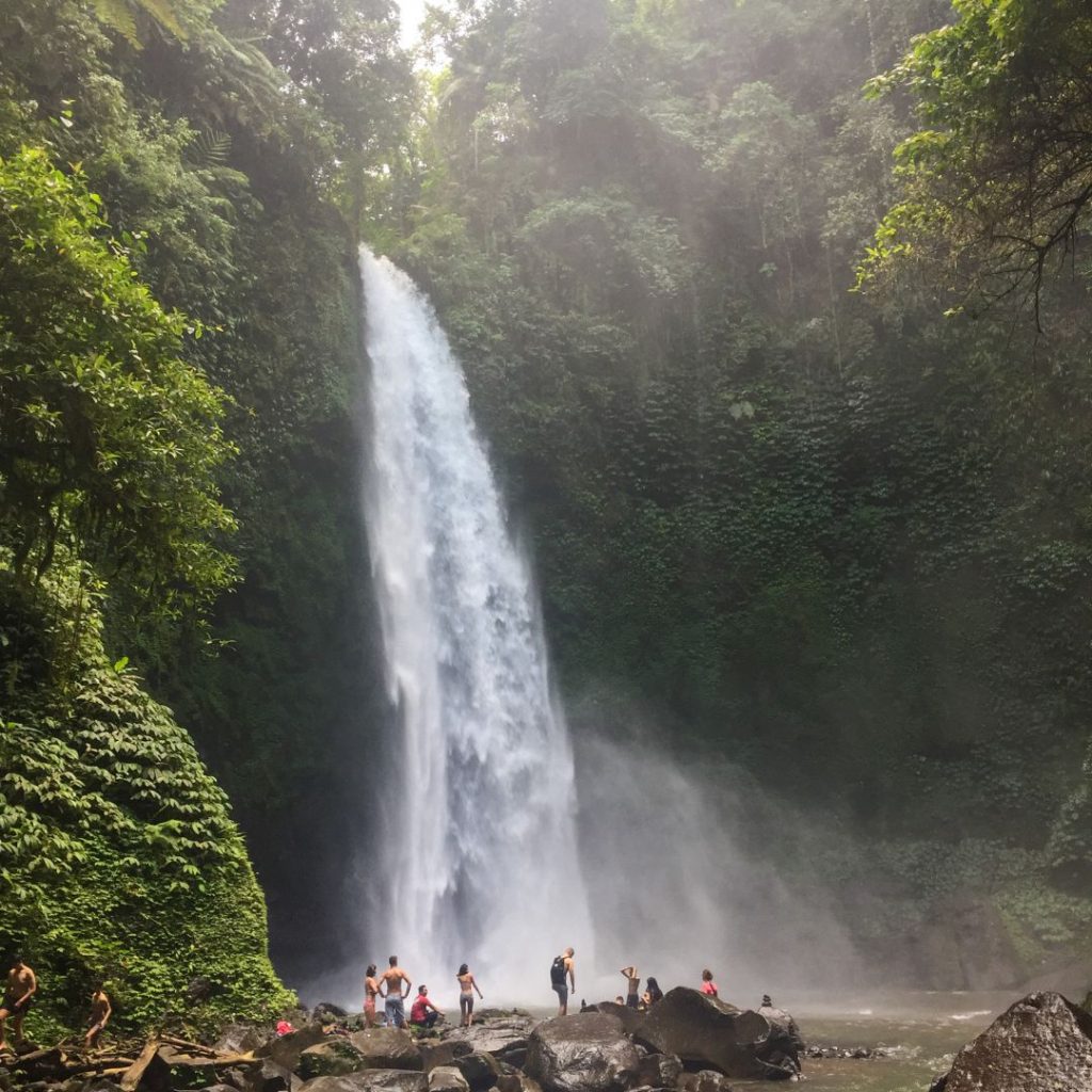 a view of the entire waterfall top to bottom as the water cascades out of the jungle and down to the pool below. There are 10 or so people standing on the boulders below watching the waterfall. There is jungle and vines surrounding the waterfall and mist rising from the pool where the water lands