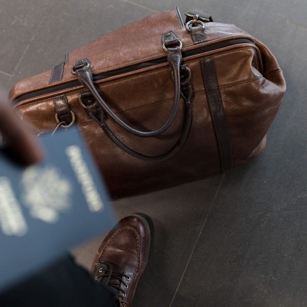 image of a brown carry-on duffle bag on the ground with a blurry image of a blue passport being held in a mans hand, ready to pack it away