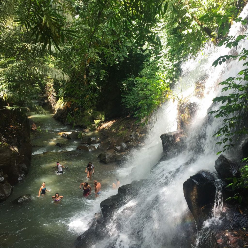 a view from the stairs looking downward to Kanto Lampo. The stepped rock formation with water gushing over the edge is to the right of the image. at the bottom of the waterfall is a number of people swimming around in the shallow water. There is lush green jungle over the canopy of the waterfall