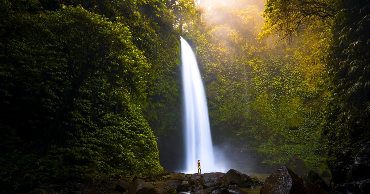 LANDSCAPE IMAGE OF WATER RUSHING OVER THE JUNGLE COVERED EDGE OF NUNGNUNG WATERFALL. THERE IS A MAN STANDING ON A ROCK AT THE BASE OF THE WATERFALL