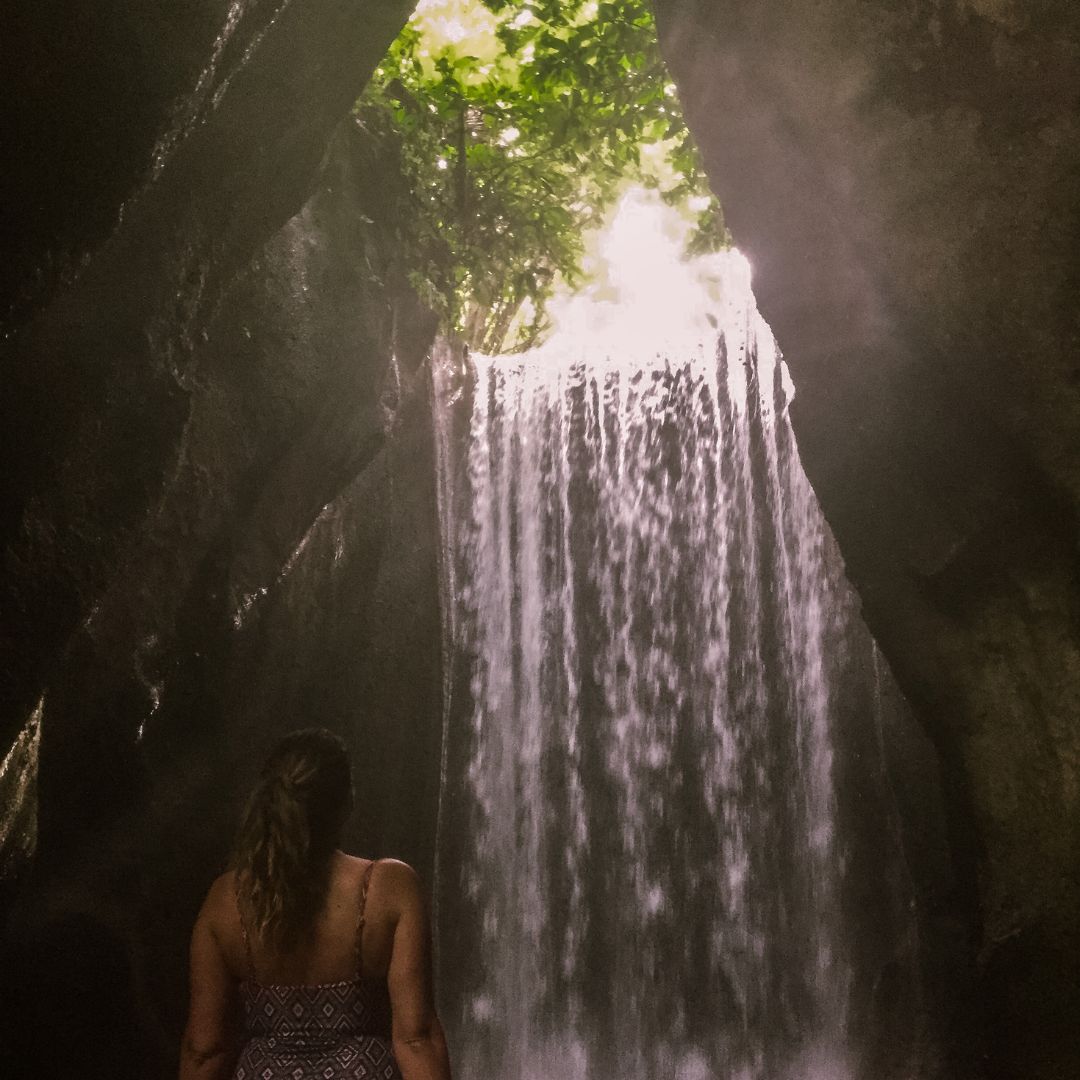 Luisa is in the waterfall cave at Tukad Cepung. she is standing in the water in her purple bathers. her hair is in a ponytail and she is looking away from the camera at the waterfall in front of her. the waterfall cascades over the edge of the rock face into the pool below