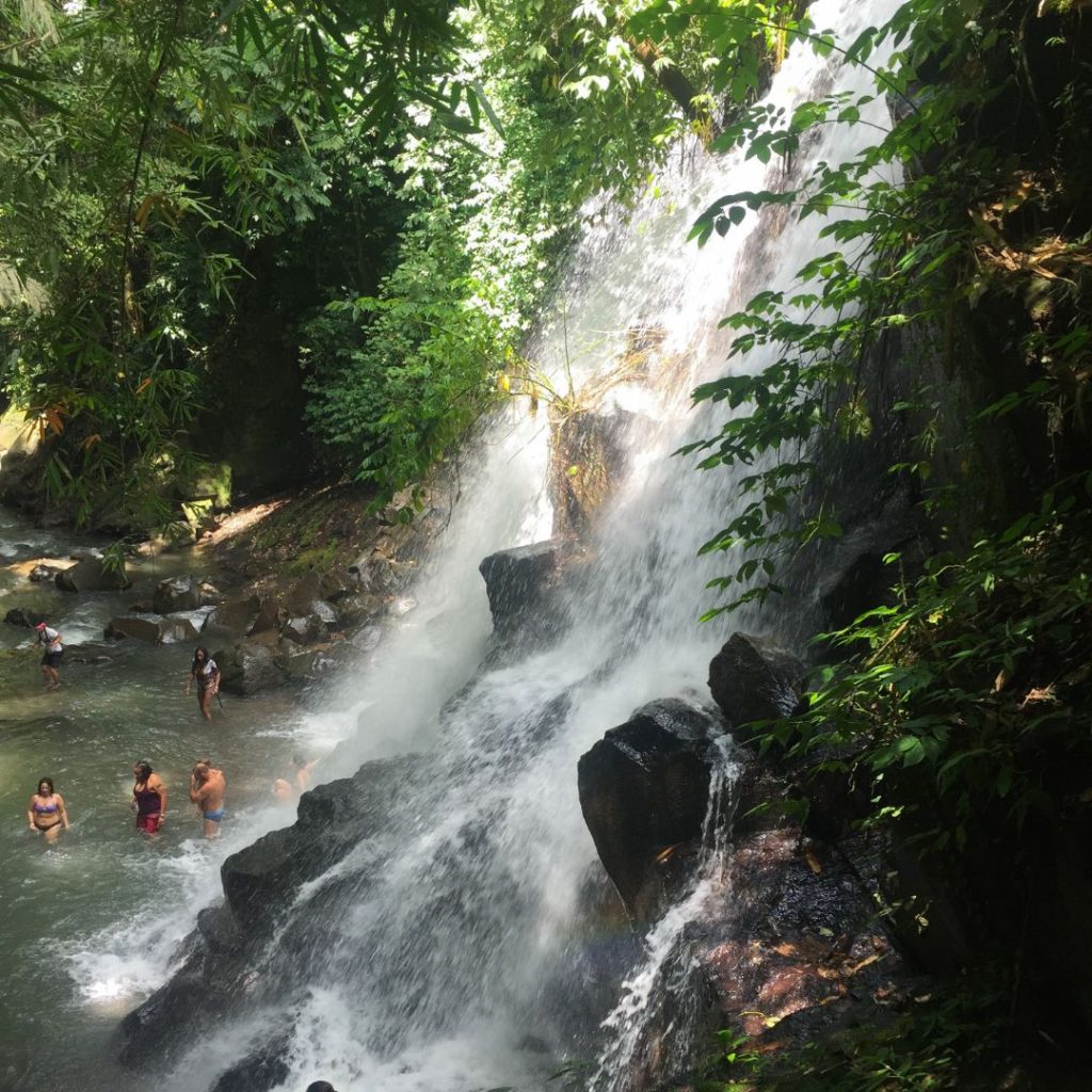 Kanto Lampo Waterfall side view from the stairs. water cascades over the edge down the step stone rock formations. people are wading in the pool at the bottom