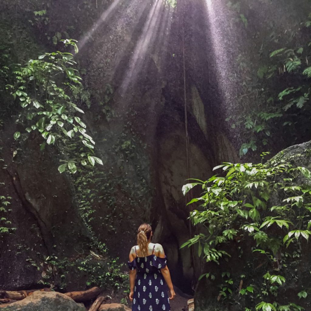 Luisa is wearing a blue and white sundress and looking away from the camera. She is looking upward toward the suns rays that are creating light beams through the canyon roof. She is surrounded by lush green jungle and high cave walls