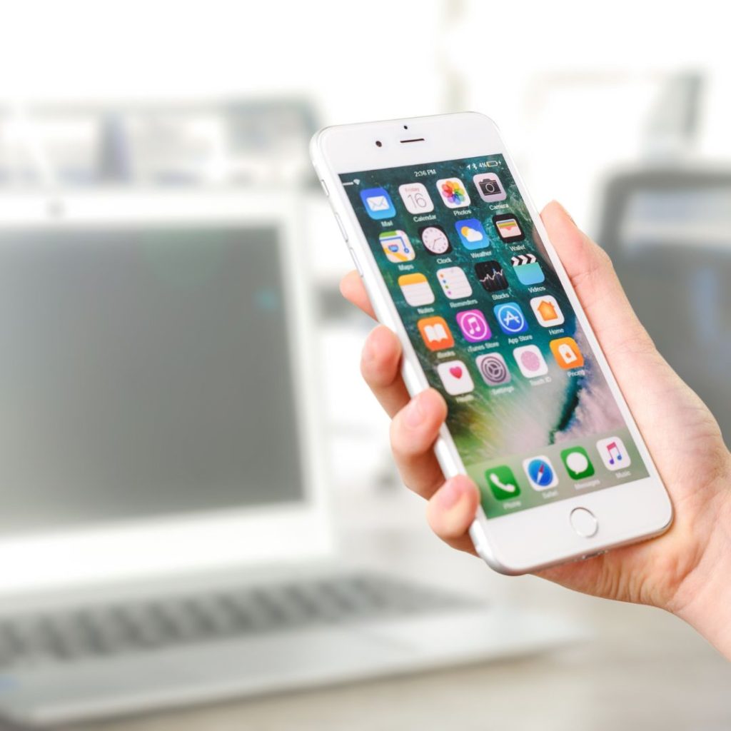 image of a hand holding a white apple iphone. In the background is a laptop.