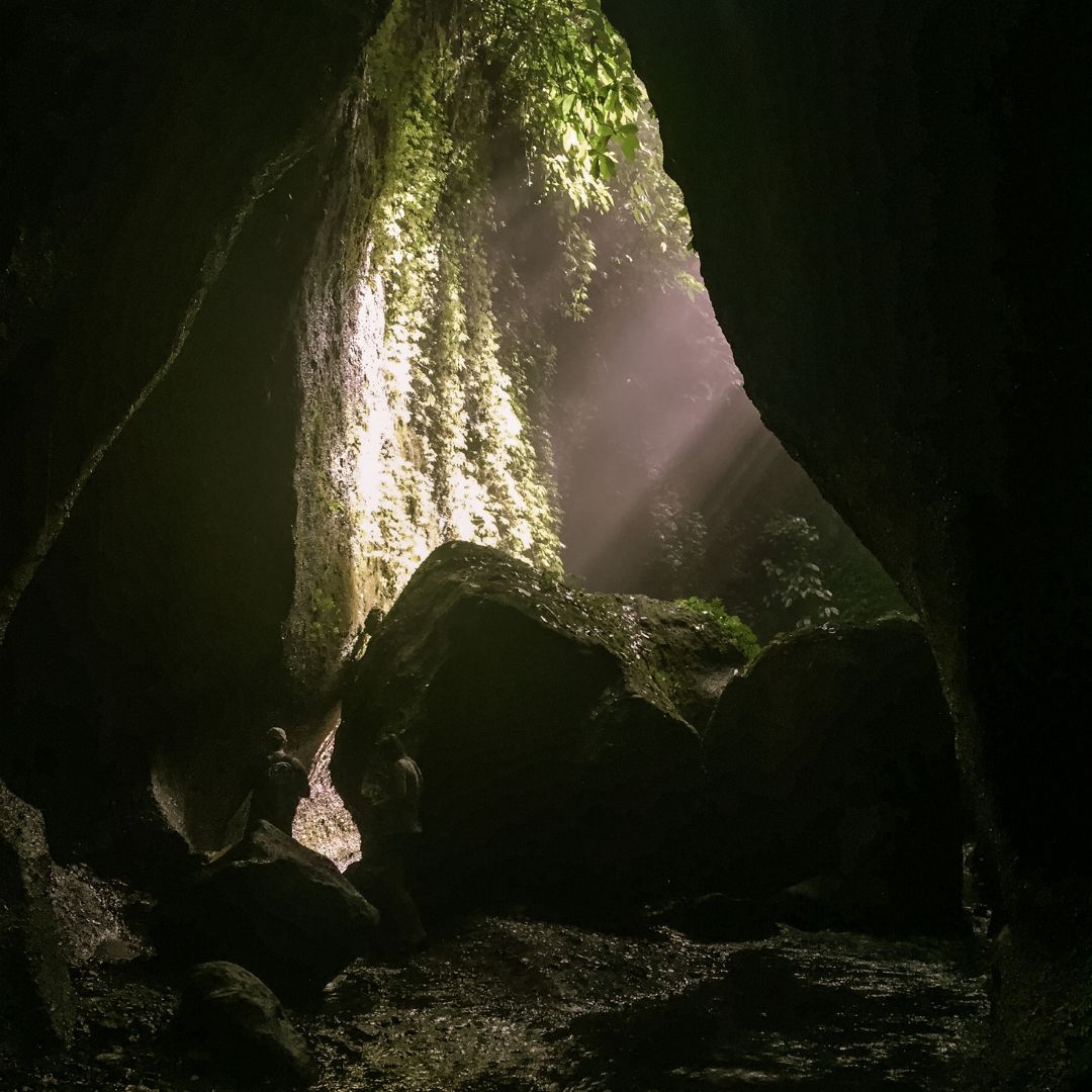 an image of the famous boulder bathed in the sun beams from the canyon above. The image is taken away from the boulder so both sides of the rock cave wall are visible but black against the sunny rock. behind the rock is lush green jungle falling down the sides of the cave wall