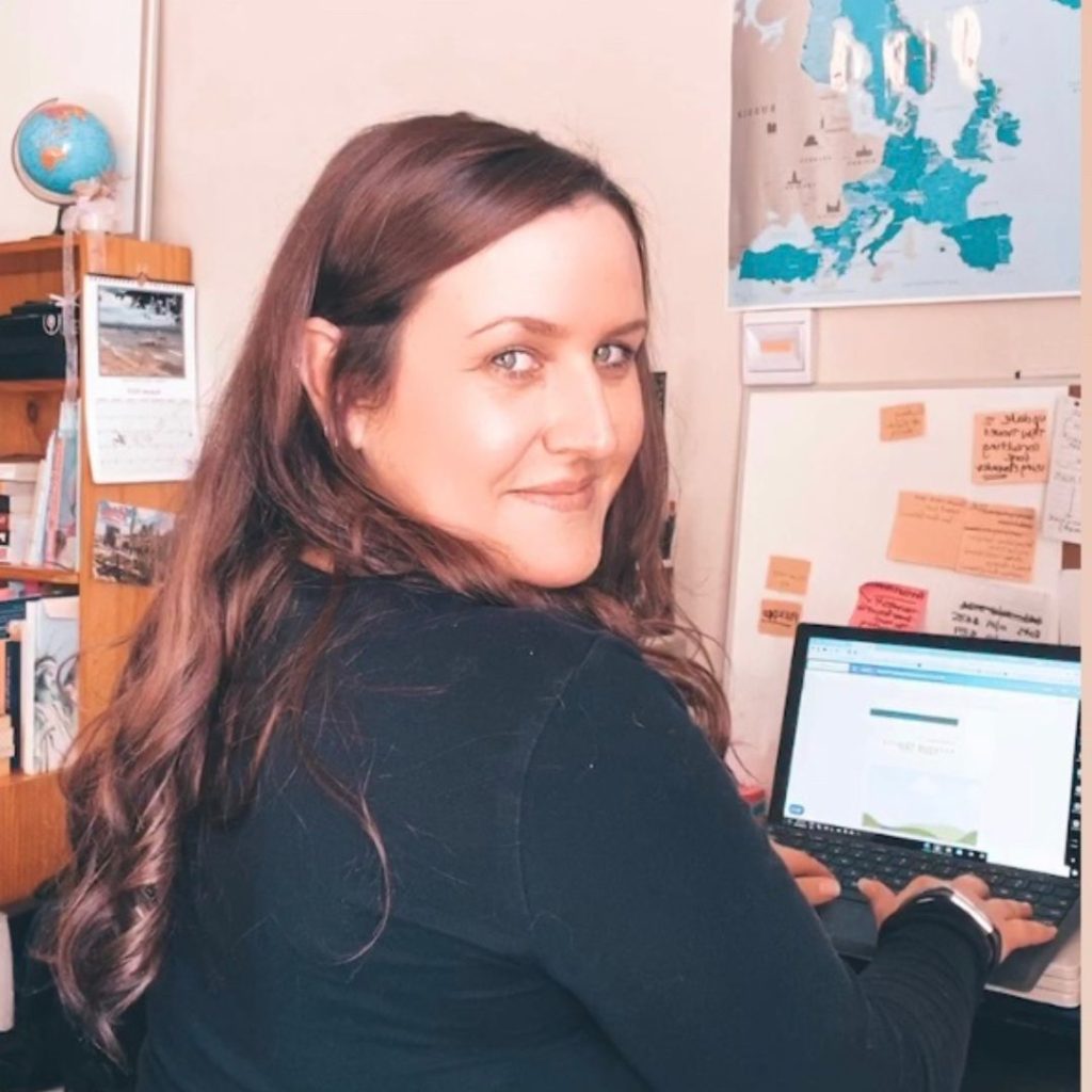 Luisa is sitting at her desk doing some travel planning on her laptop while looking over her right should back toward the camera