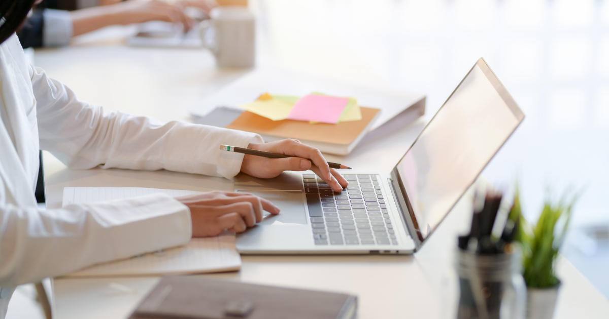 woman sits at a desk typing on her laptop. her desk is covered in papers, notebooks and a plant