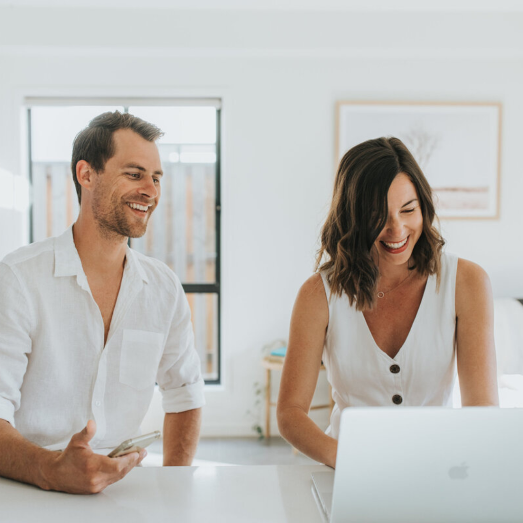 Kev to the left and Jade to the right both dressed in white, sit at a bench. They are the creators of the affiliate marketing course for beginners. Jade is working on her laptop and Kev is holding his mobile phone. They are both smiling and laughing